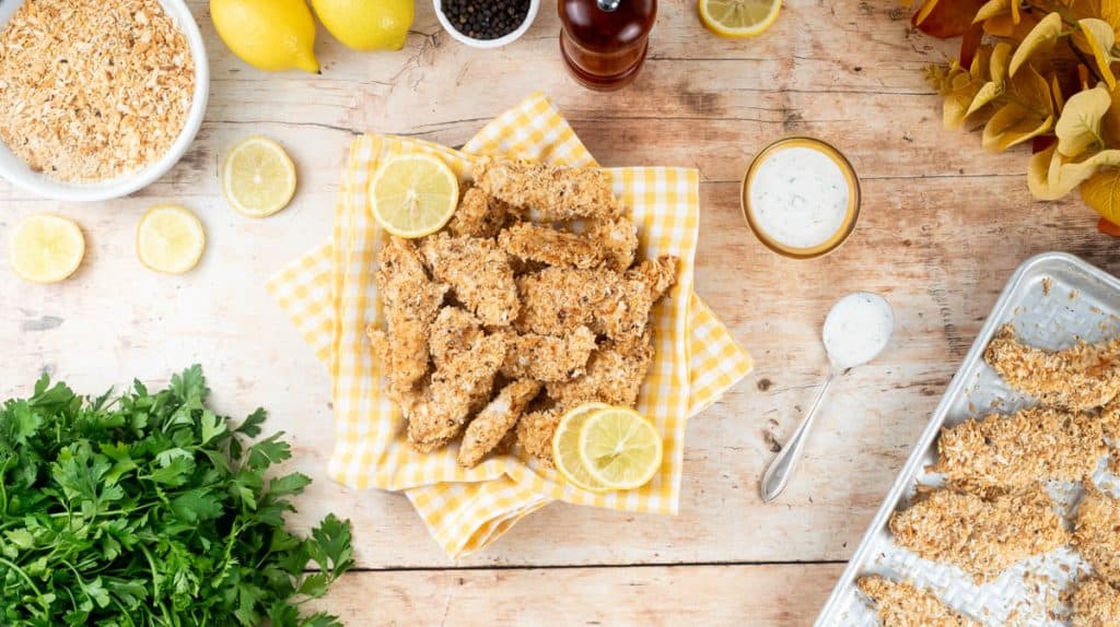 An overhead view of a pile of lemon pepper baked chicken tenders laid on a yellow napkin. They are surrounded by decorative items, like fresh parsley, lemon slices and extra breadcrumbs.