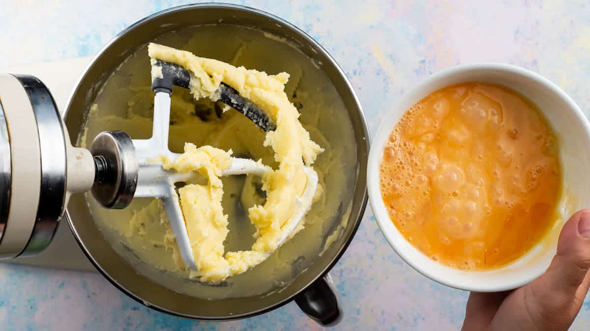 A hand is pouring whisked egg into the butter-sugar mixture.