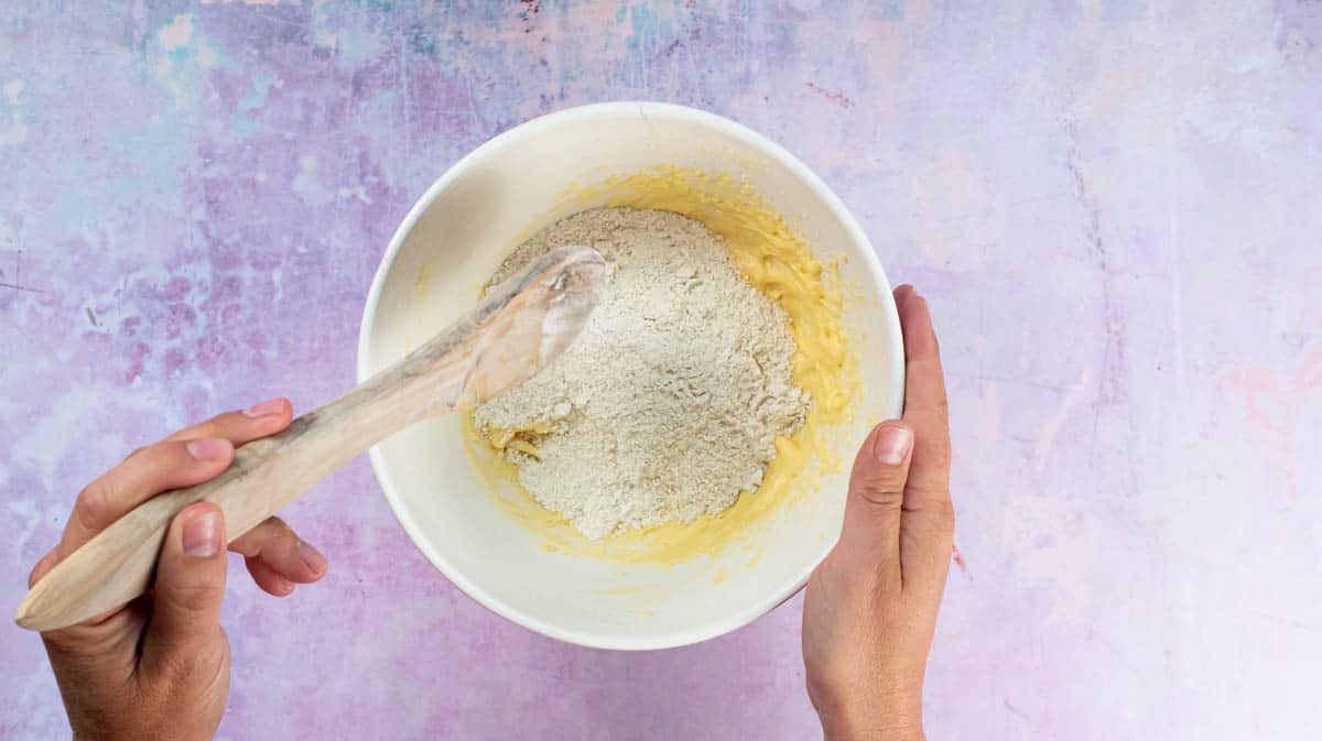 A hand is holding a wooden spoon and is about the stir the flour and ground almond mixture into the wet ingredients.