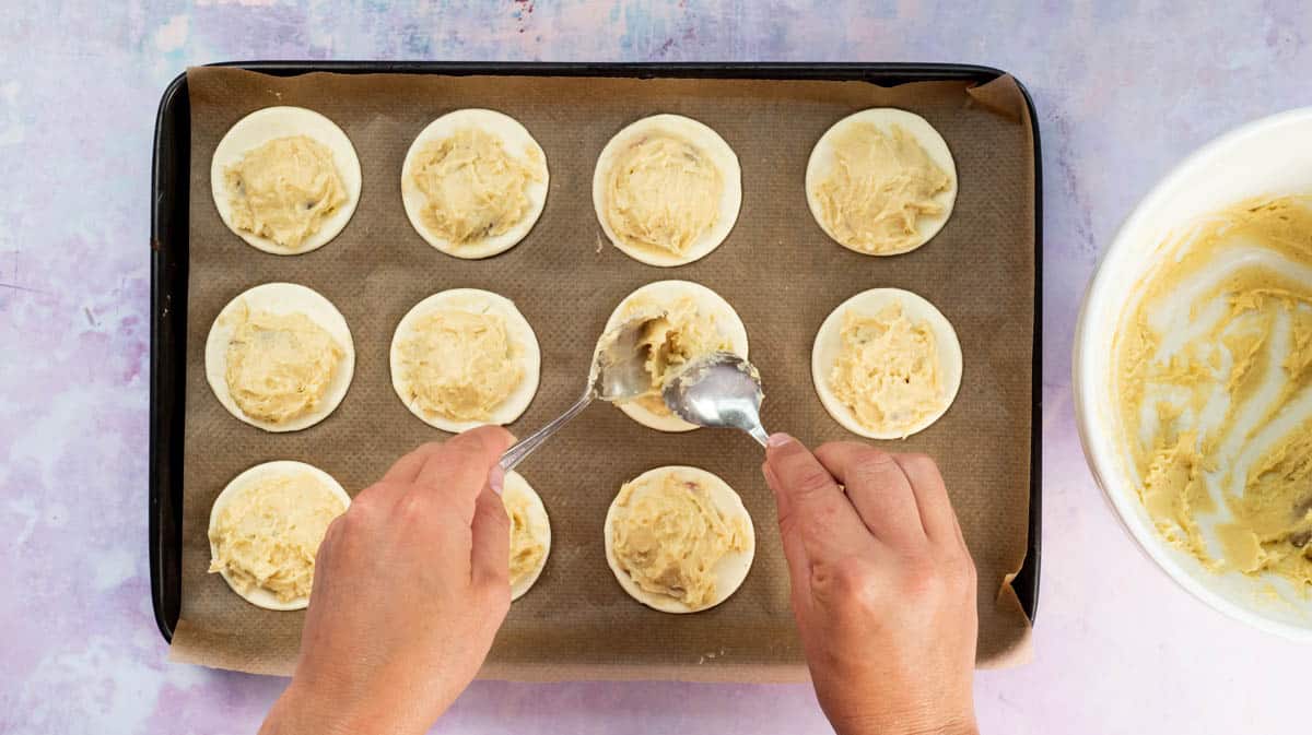 Two hands are using spoons to cover the jam on the puff pastry circles in Frangipane.
