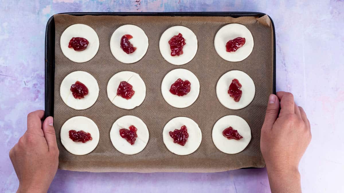 Two hands are holding a lined baking tray with twelve circles of puff pastry laid out on it. Each one has a spoonful of jam in the middle.