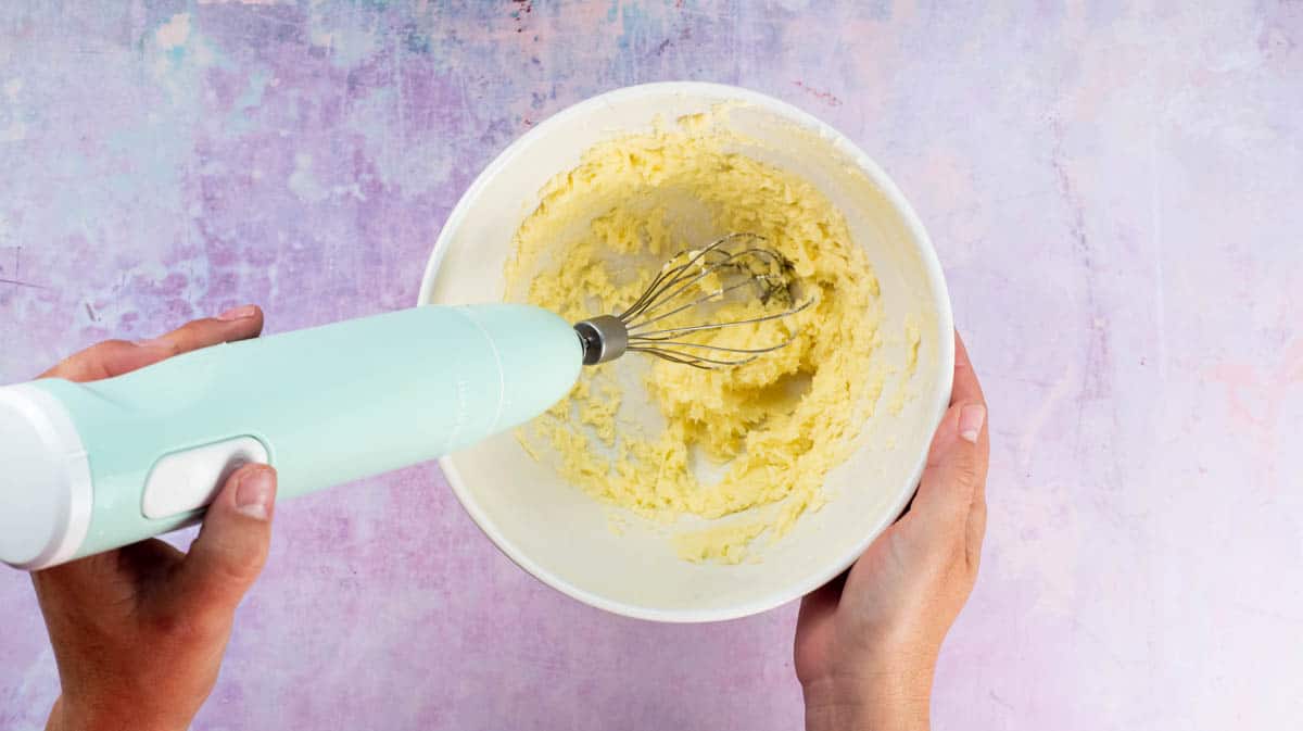 A hand is using an electric whisk to mix the butter, almond essence and sugar together.