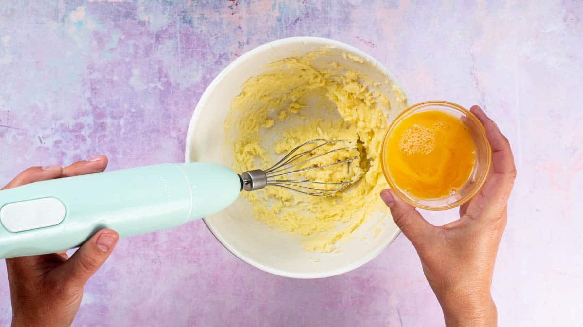 A hand is preparing to pour whisked egg into the butter sugar mixture.