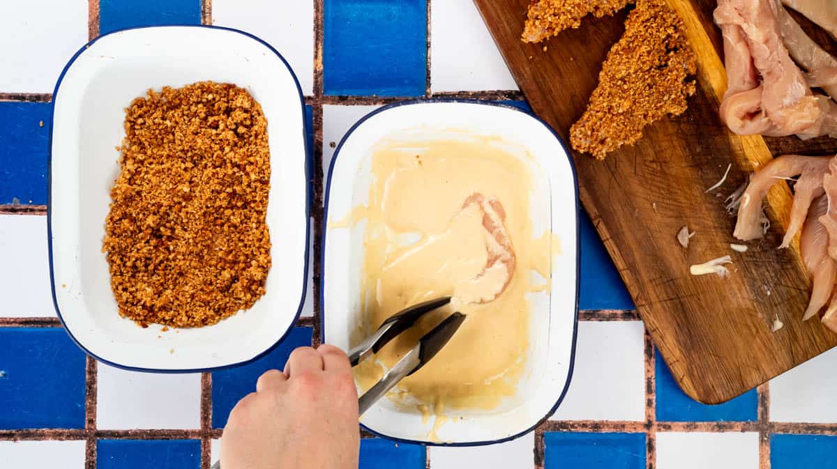 A hand is using tongs to turn a piece of chicken over in the mayonnaise batter.