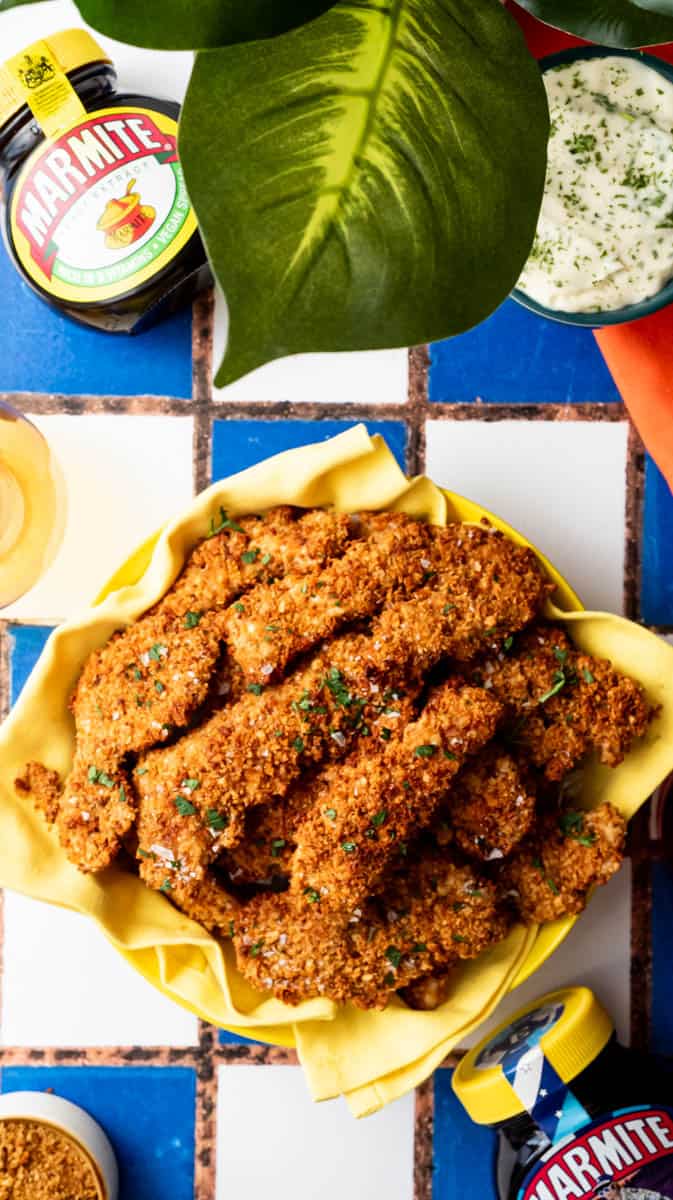 A yellow bowl filled with Marmite chicken dippers surrounded by decorative items and ingredients.