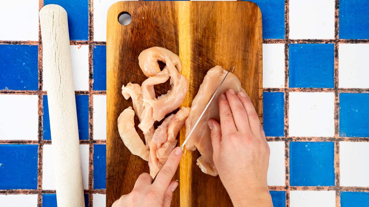 I hand is using sharp knife to slice flatten chicken breast into pieces.