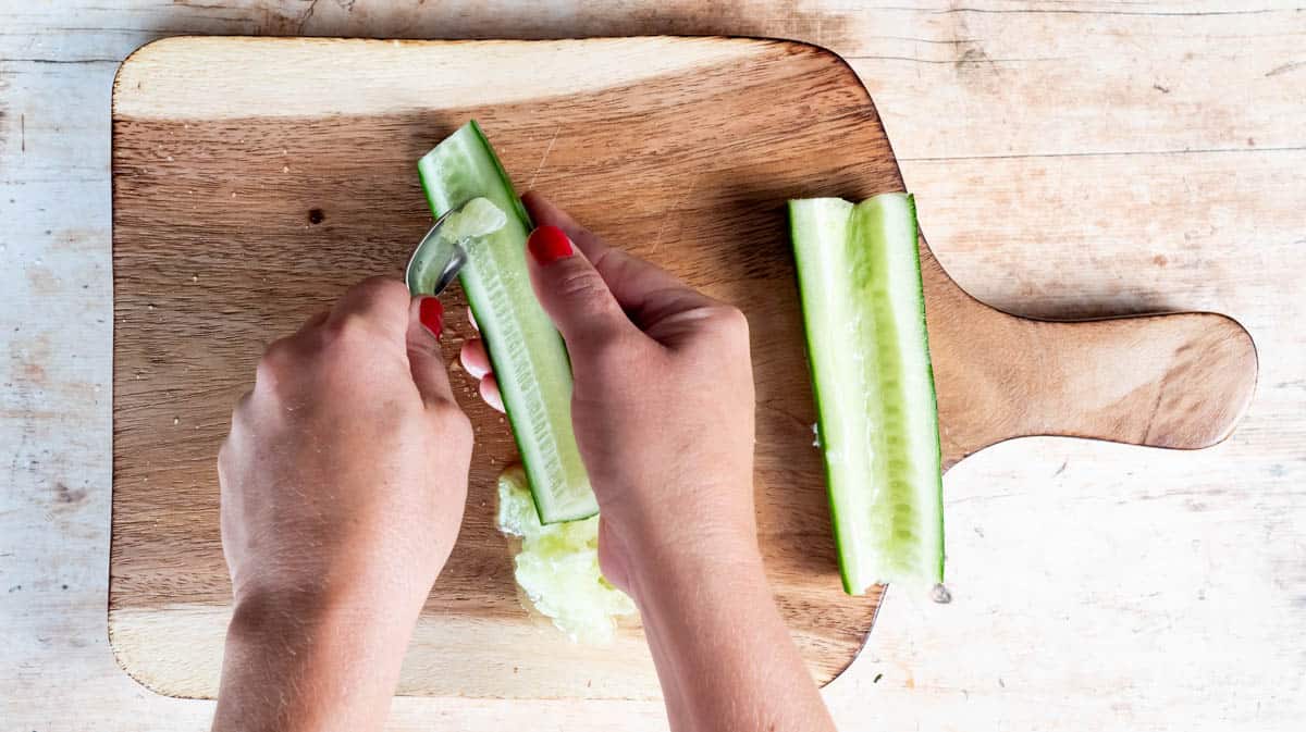 A hand is using a spoon to scrape the seeds from the middle of a cut cucumber full stop