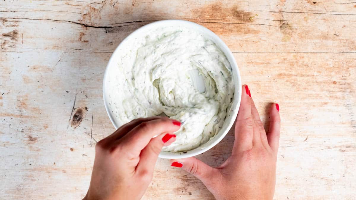 A hand is using a spoon to mix together the ingredients of the Raita sauce inside a white bowl.