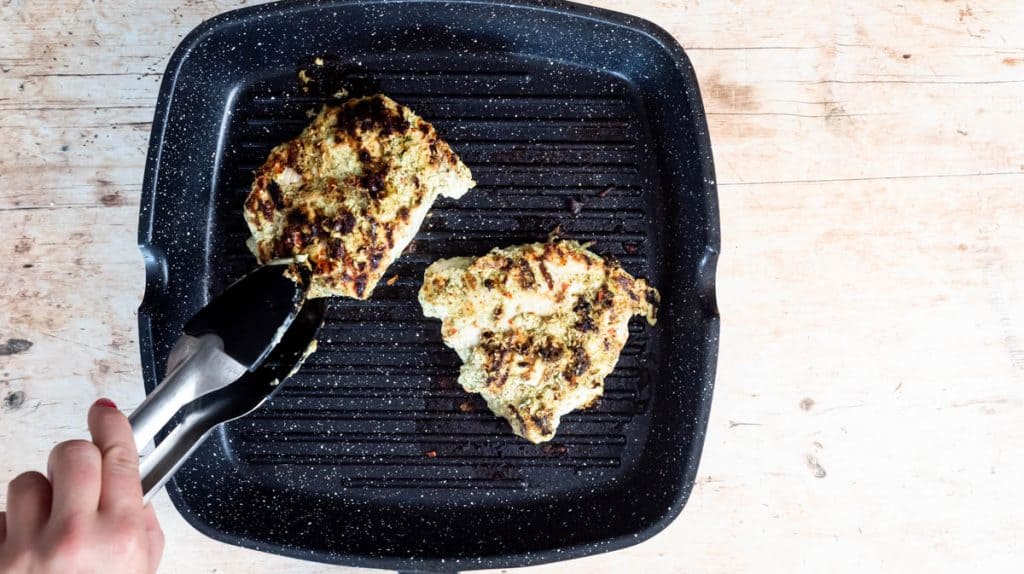 A hand is using tongs to turn chicken breast inside a grill pan. Both chicken breastsare browned on one side.