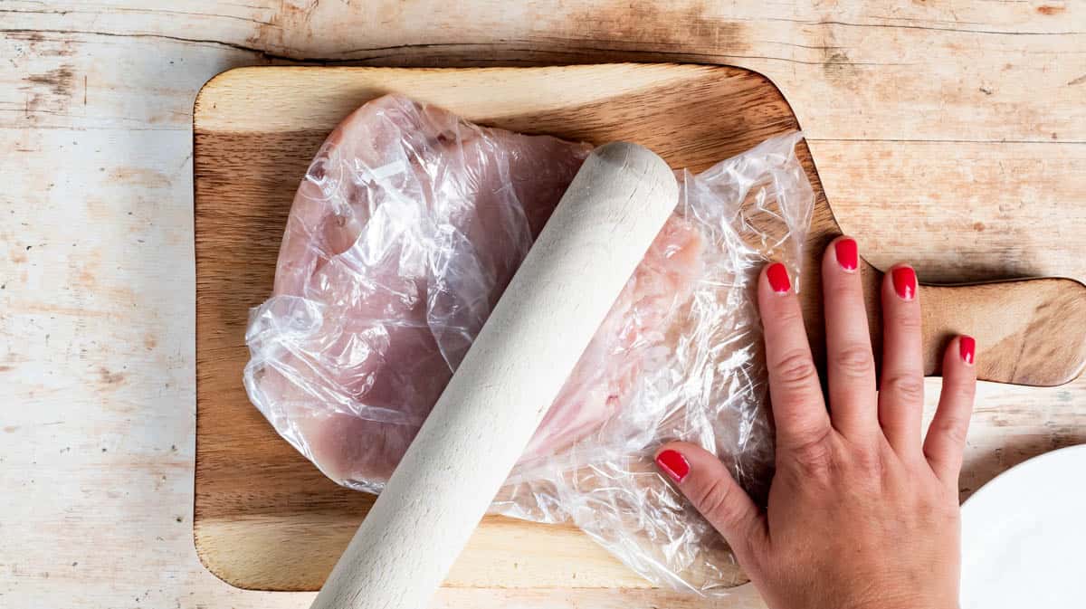 A hand is using a rolling pin to flatten chicken that's inside a food bag.