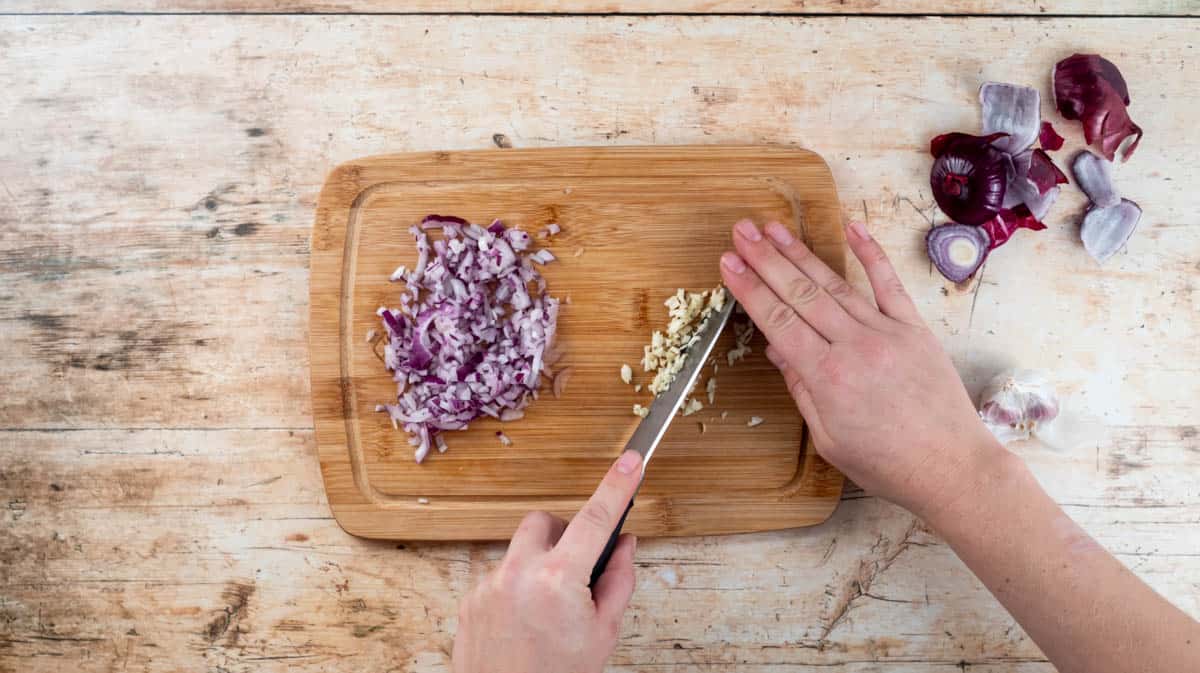 A hand is using a knife to finely chop the garlic on a wooden board that also has finely chopped red onion on it.