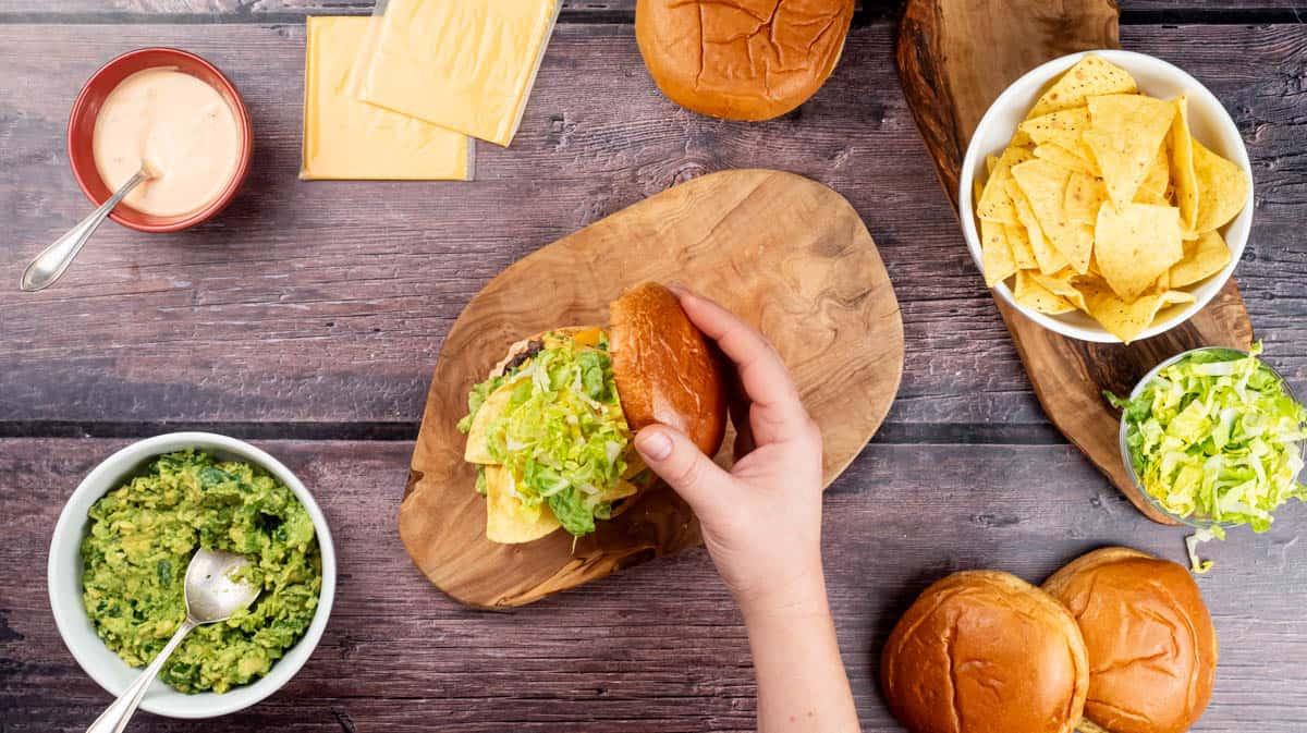 A hand is placing the top half of a burger bun on top of a filled nacho burger.