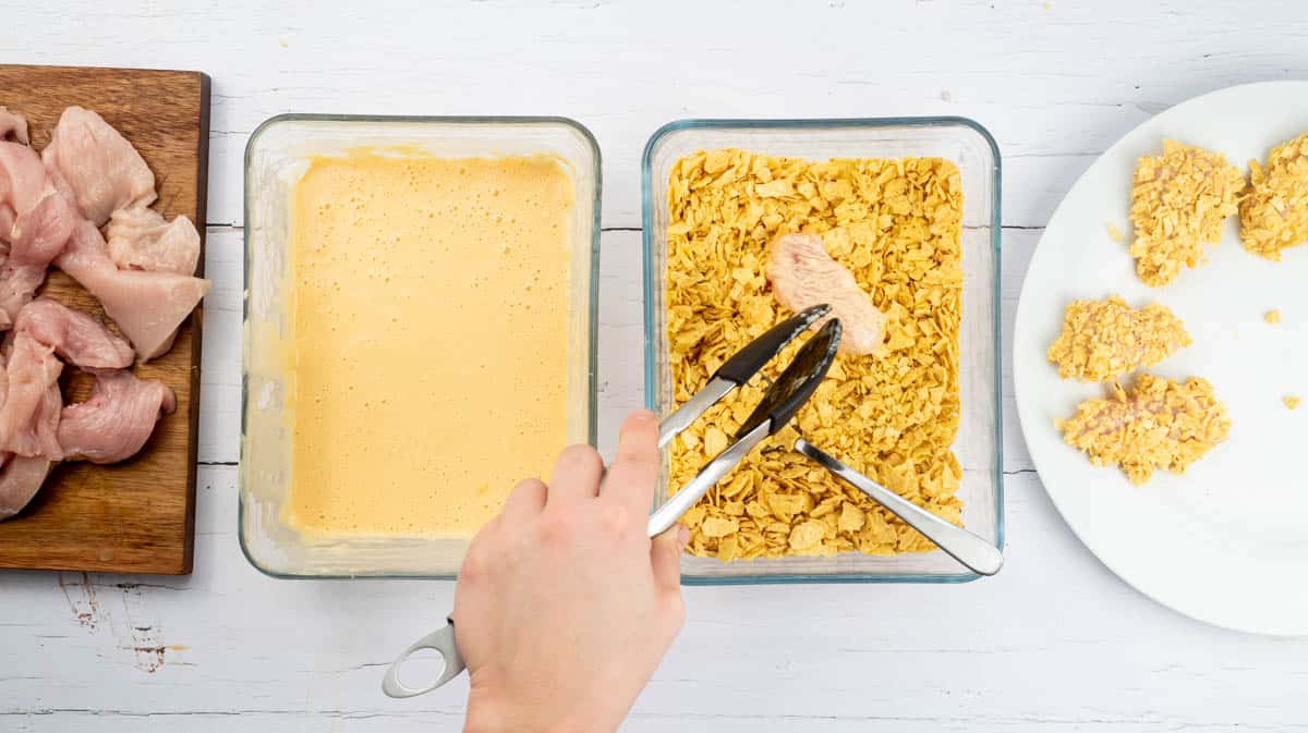 A hand is using tongs to transfer a chicken piece coated in batter into a bowl of crushed nachos.