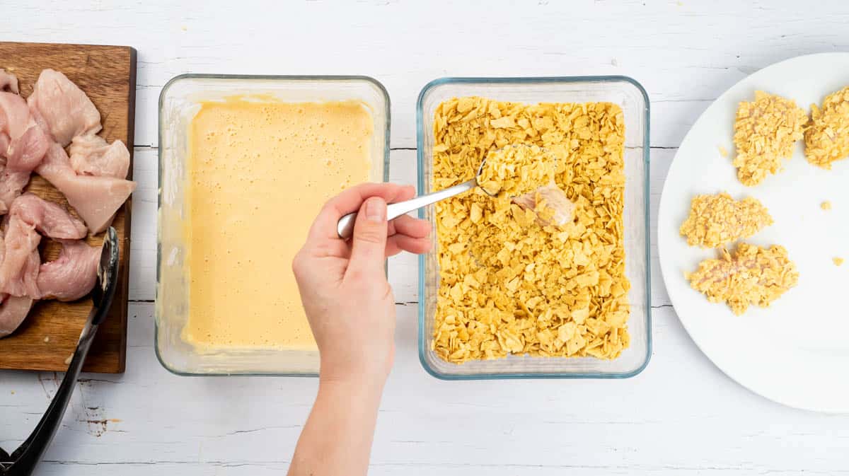A hand is using a spoon to cover the battered chicken piece with crushed nachos.