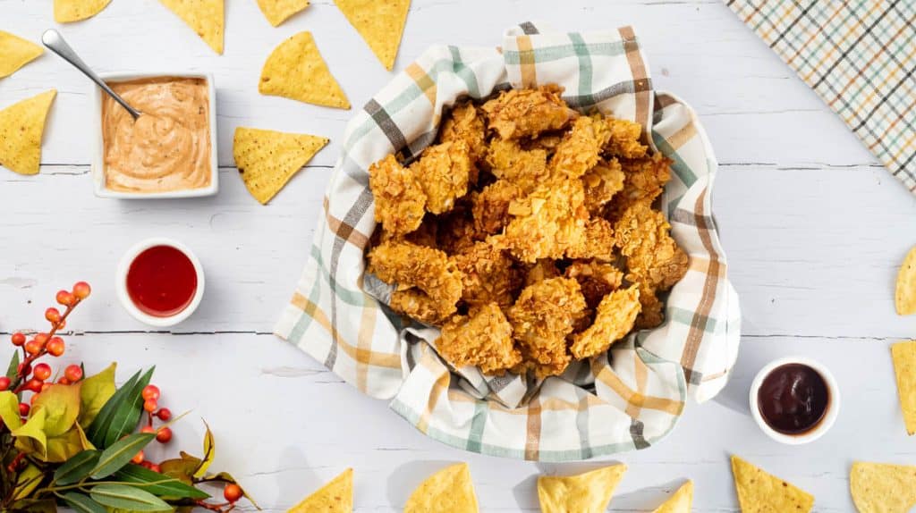 An overhead view of a bowl of nacho chicken bites surrounded by dips and nachos.