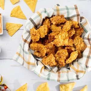 An overhead view of a bowl of nacho chicken bites surrounded by dips and nachos.
