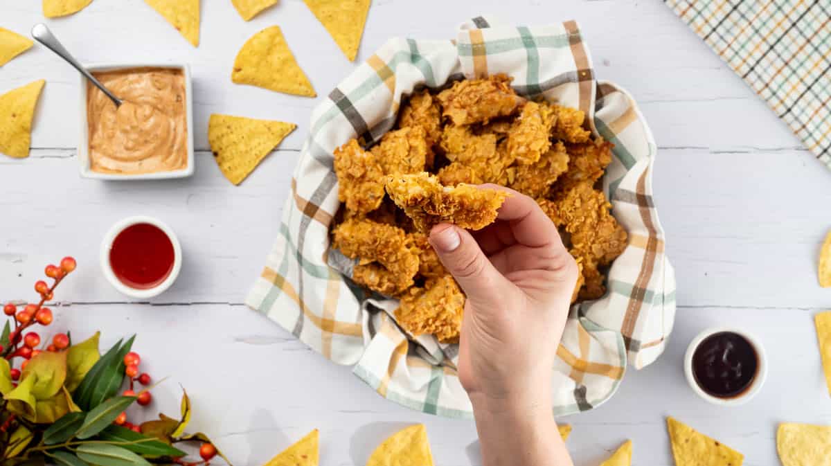 A hand is holding a nacho chicken bite up to the camera. There is a bowl with a pile of others in the background along with dips and nachos.