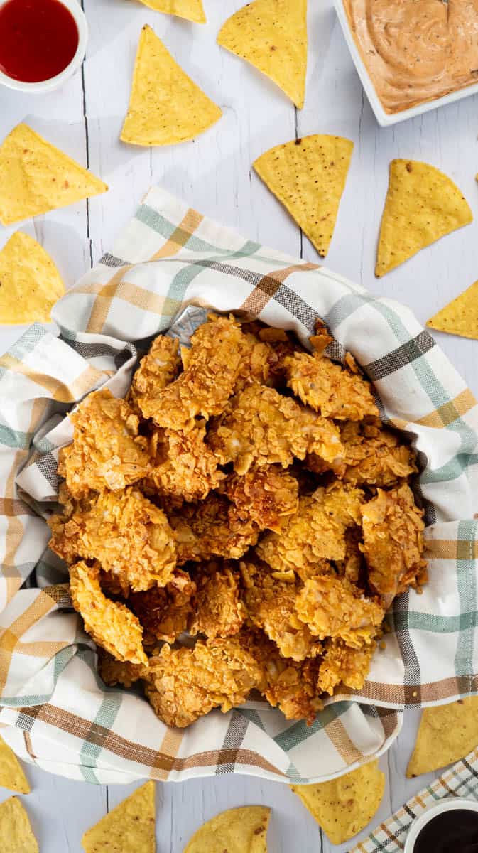 A lined tin with a pile of nacho chicken bites inside. There are dips and nachos around the tin in a random pattern.