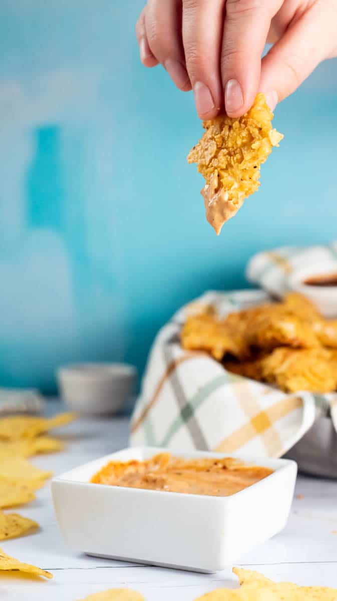 A hand is holding a nacho coated chicken bite that has been dipped in a sauce.