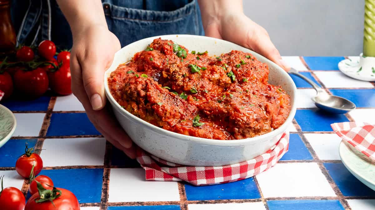 Two hands are cupping a bowl filled with beef meatballs covered in a tomato sauce.