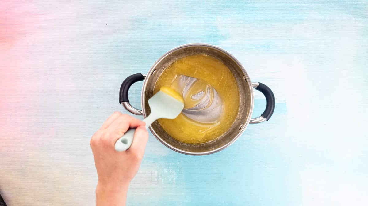 A hand is using a blue spatula to mix butter, caster sugar and golden syrup as it melts inside a saucepan.