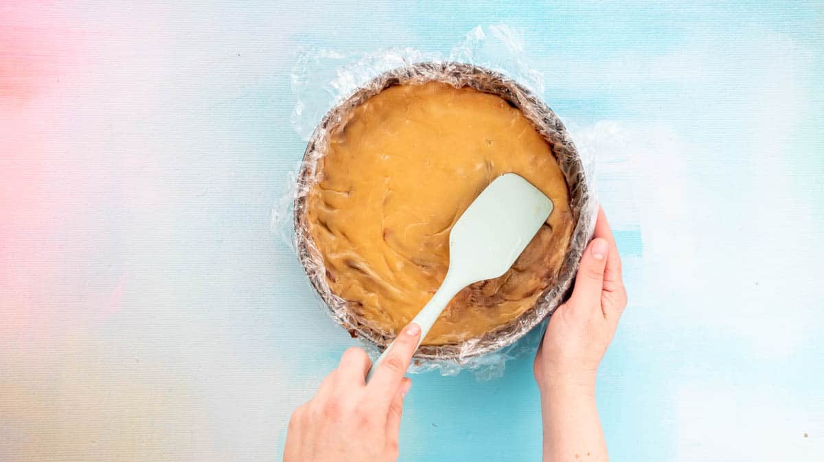 A hand is using a blue spatula to spread the caramel layer over the top of a Nutella layer.