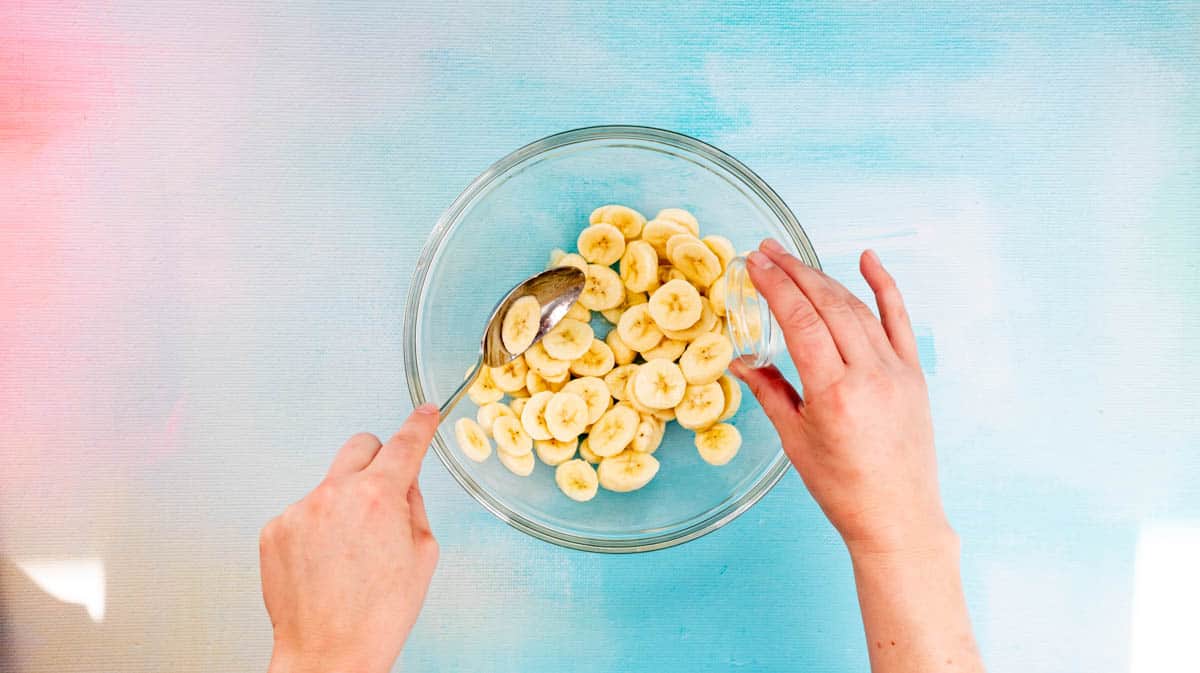 A hand is pouring lemon juice over sliced bananas inside a glass bowl.