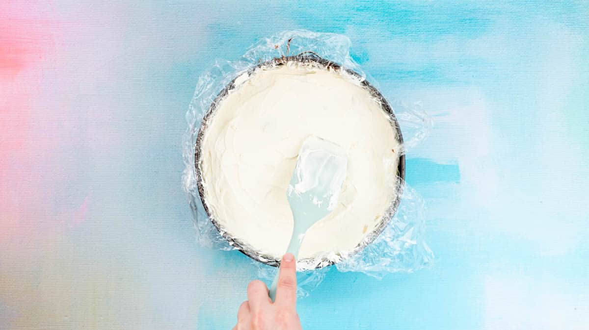 A hand is using a spatula to spread whipped cream over the top of the Banoffee pie inside a cake tin.