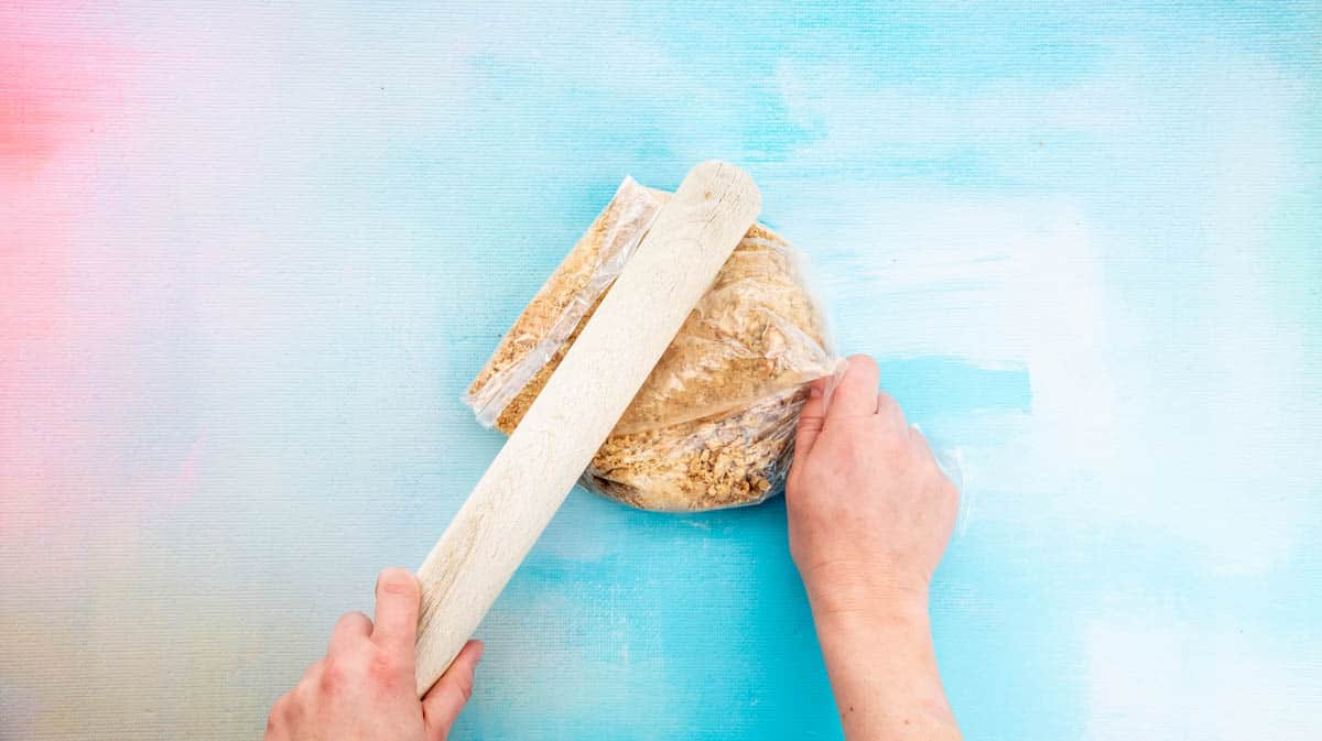 A hand is using a rolling pin to crush digestive biscuits into crumbs batter inside a food bag.