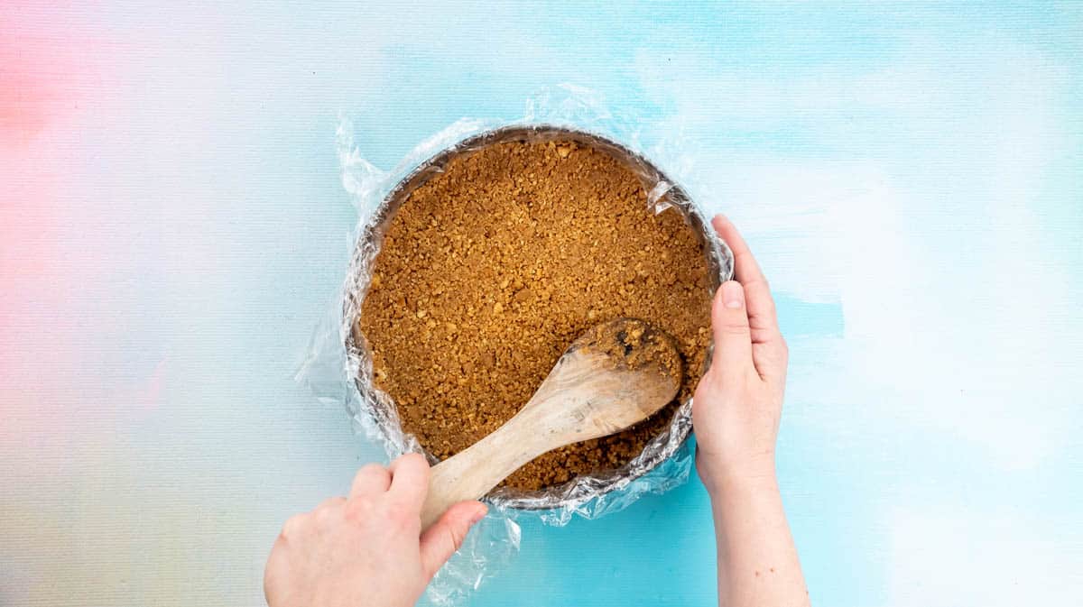 .A hand is using a wooden spoon to press the biscuit crumbs down firmly inside a cake tin.