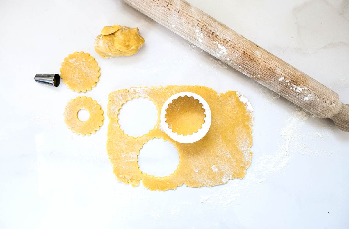 Party rings biscuit dough rolled out, with a cookie cutter on top, next to pre-cut cookie dough and an example of one with a hole cut out using a piping bag nozzle.