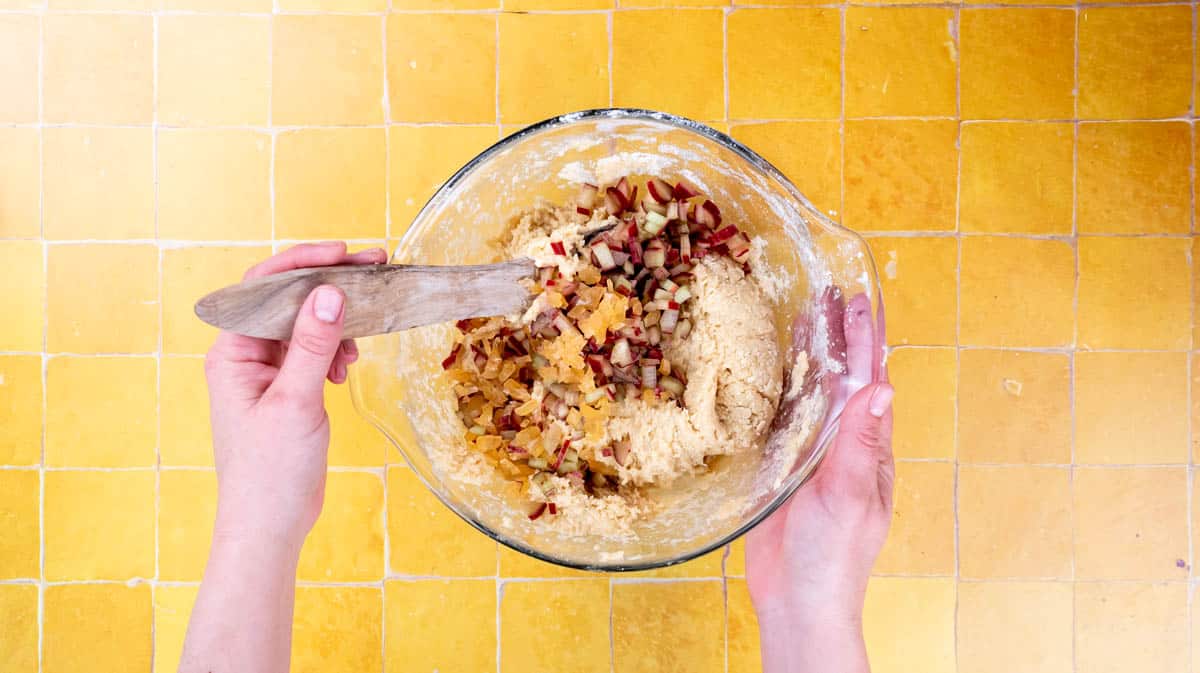 A hand is using a wooden spoon to mix diced rhubarb and ginger into the scone dough.