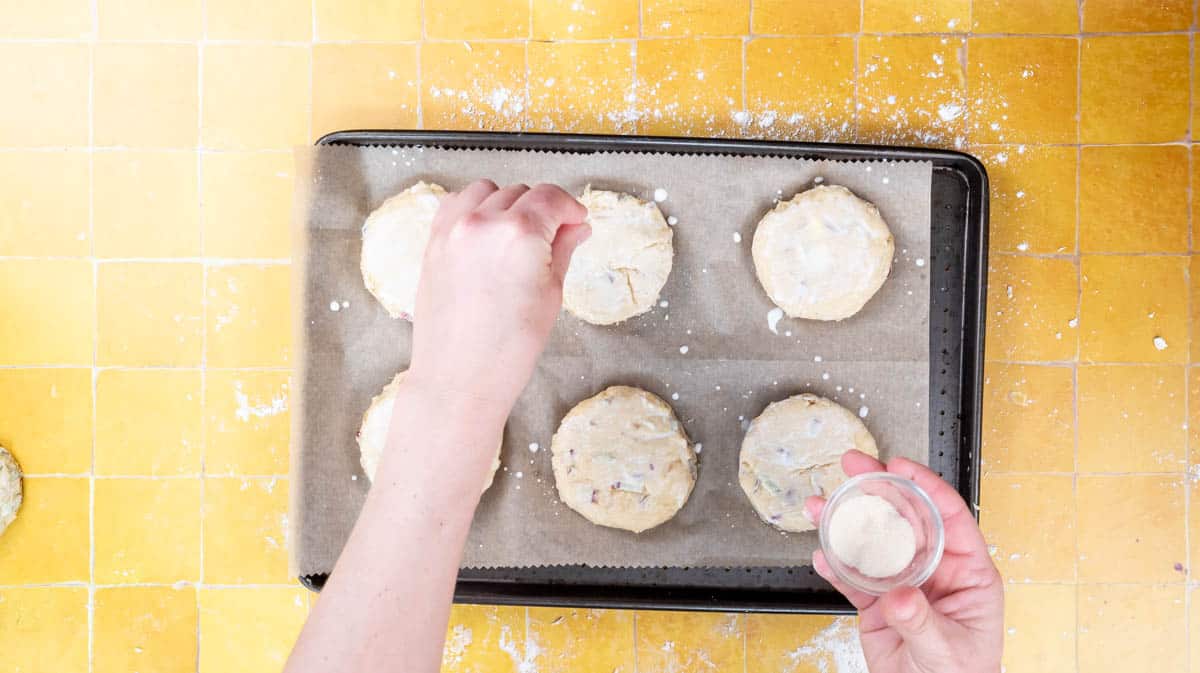 A hand is sprinkling caster sugar onto the top of the unbaked scones.