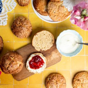 A sliced rhubarb scones with clotted cream and jam spread on one half. Surrounded by more scones and ingredients.