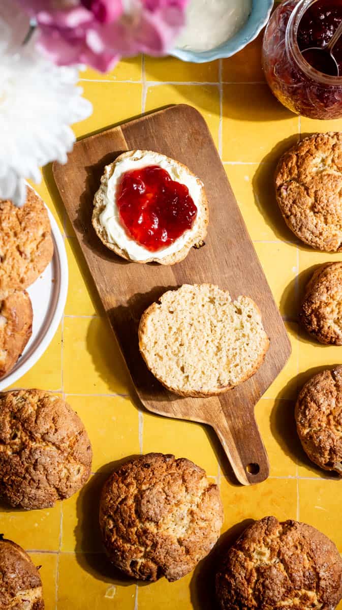 A sliced rhubarb scone on a wooden chopping board with one half covered in cream and jam.