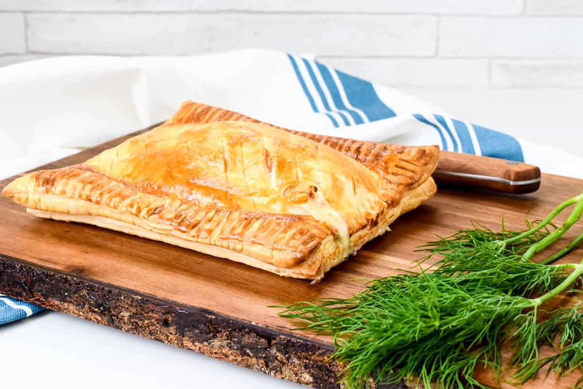 A golden-brown baked salmon parcel on a wooden board next to a bunch of fresh dill.