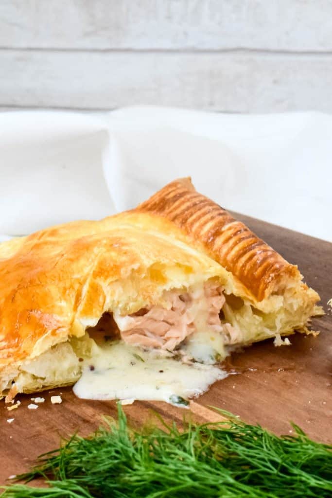 Salmon En Croute on a wooden board, cut in half with the dill sauce pouring out.
