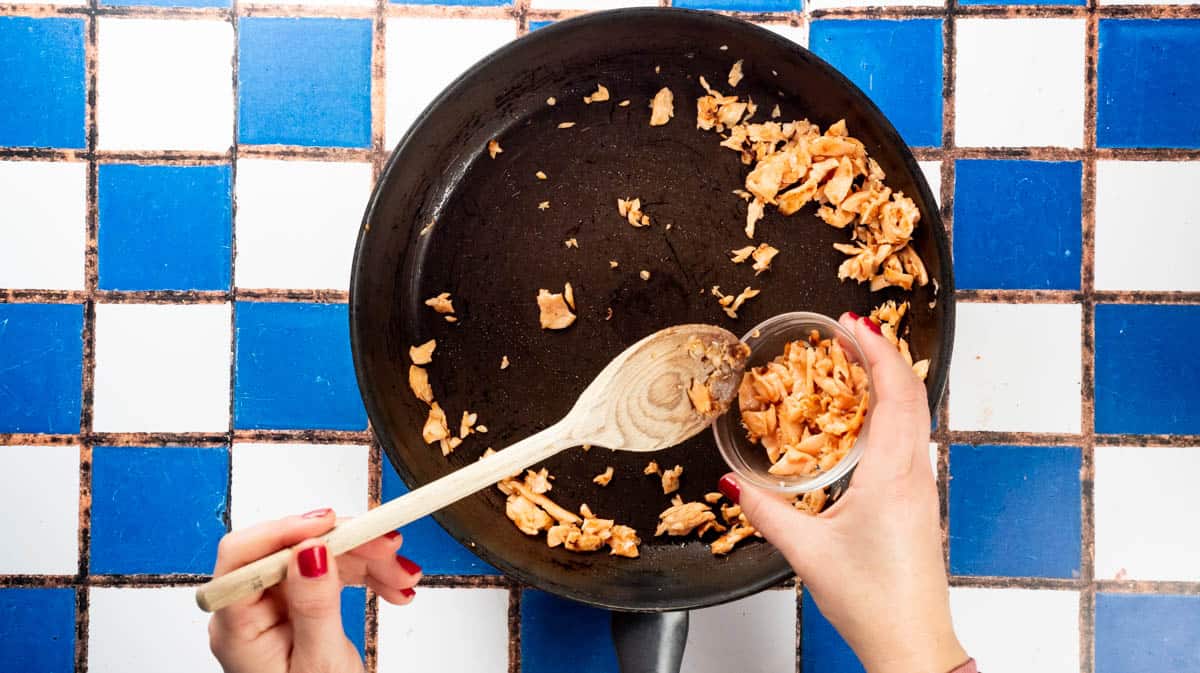 A hand is using a wooden spoon to collect some of the salmon mixture and place it in a glass bowl.