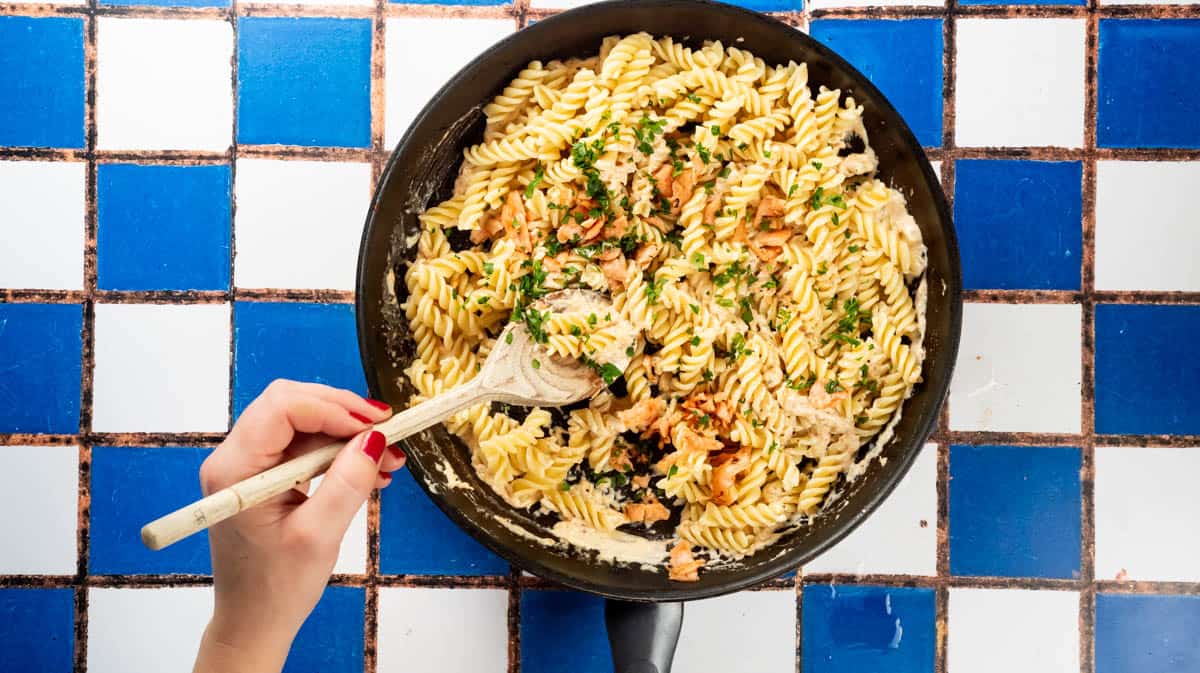 A hand is using a wooden spoon to mix in reserved salmon and parsley to the pasta mixture.