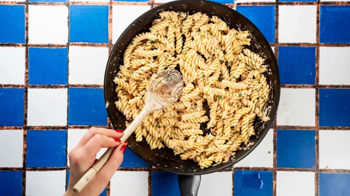 A hand is using a wooden spoon to mix cooked pasta with the smoked salmon sauce.