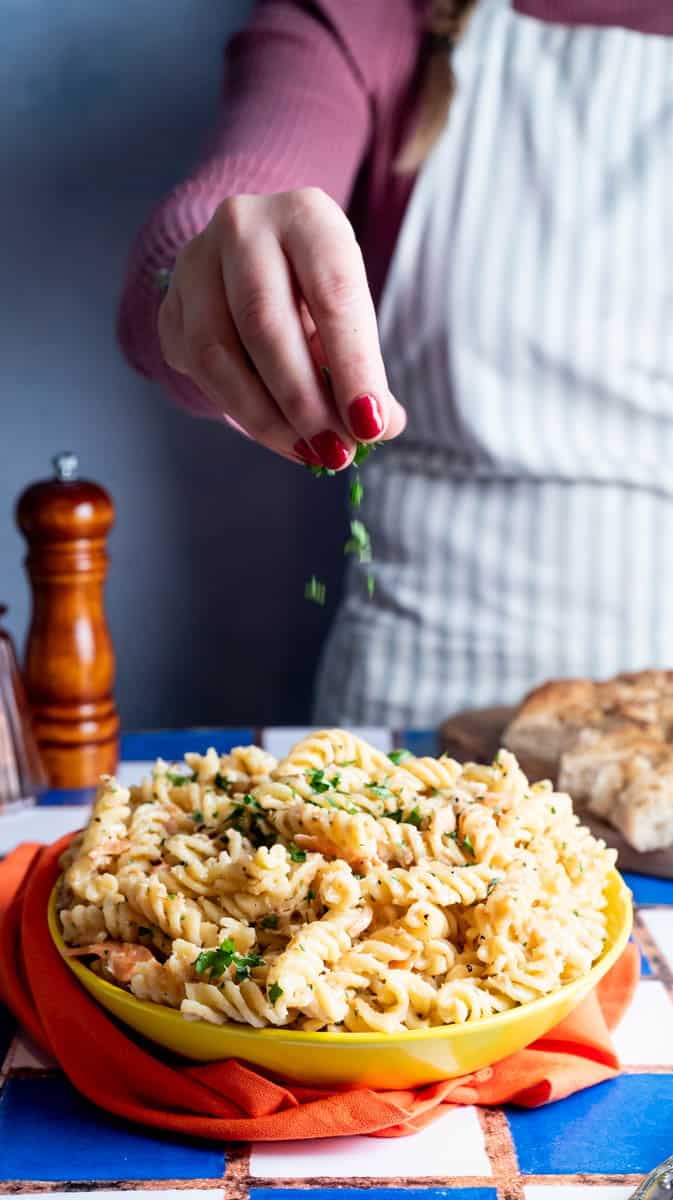 A person is sprinkling fresh parsley over a bowl of salmon pasta.