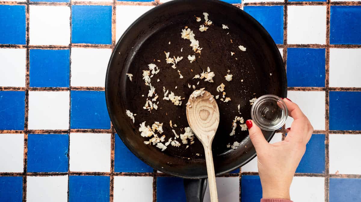 A hand is pouring white wine into a frying pan with sauteed onion inside.
