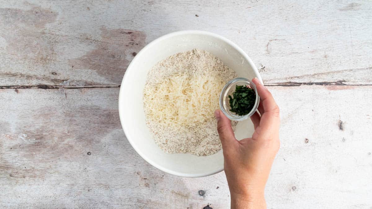 A hand is tipping parsley into a bowl containing a flour-butter mixture and parmesan cheese.