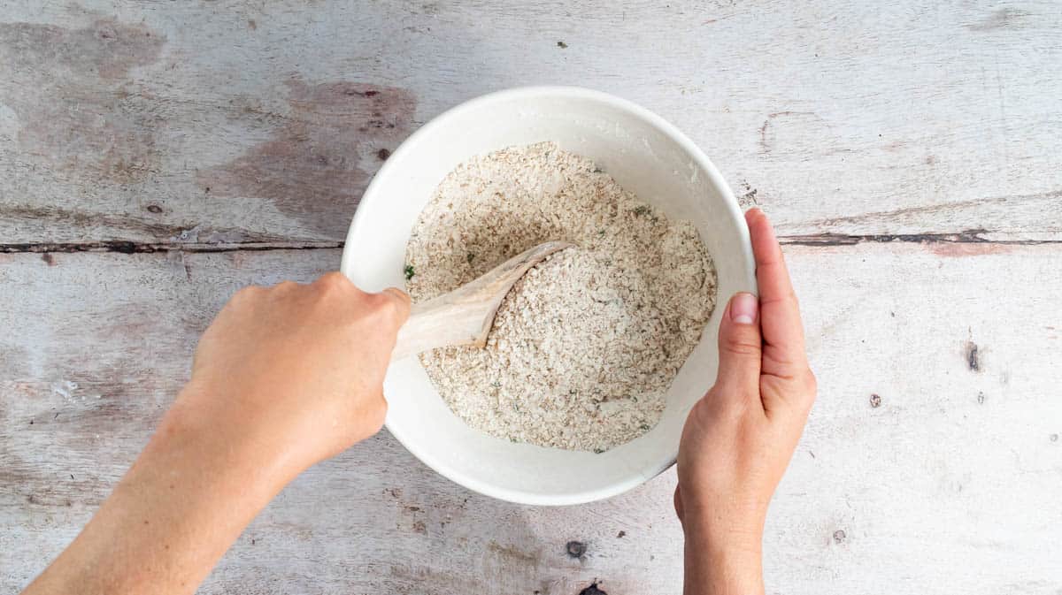 A hand is using a wooden spoon to mix the dry ingredients of a soda bread loaf together in a bowl.
