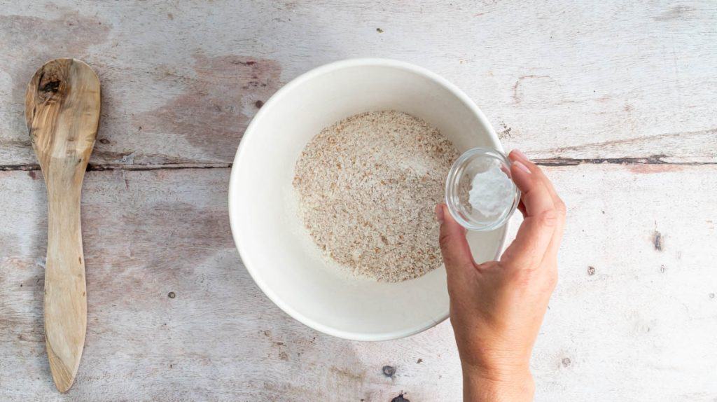 A hand is adding bicarbonate of soda to a bowl with flour inside.