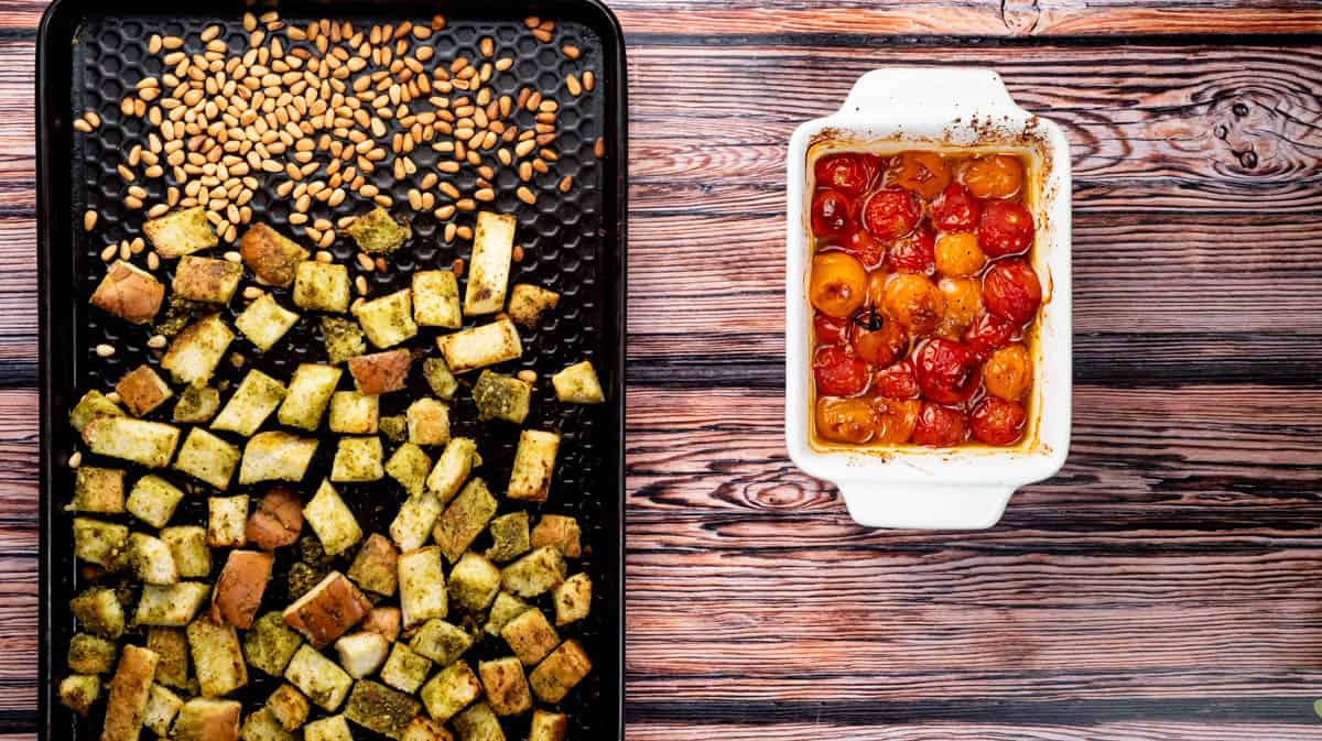 A baking tray with toasted pine nuts and bread cubes on it next to a bowl of roasted cherry tomatoes.
