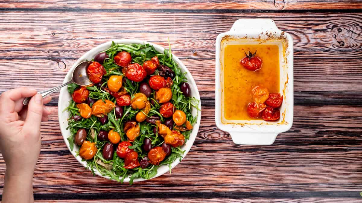 A hand is using a spoon to place roasted tomatoes into a bowl with rocket leaves and olives.
