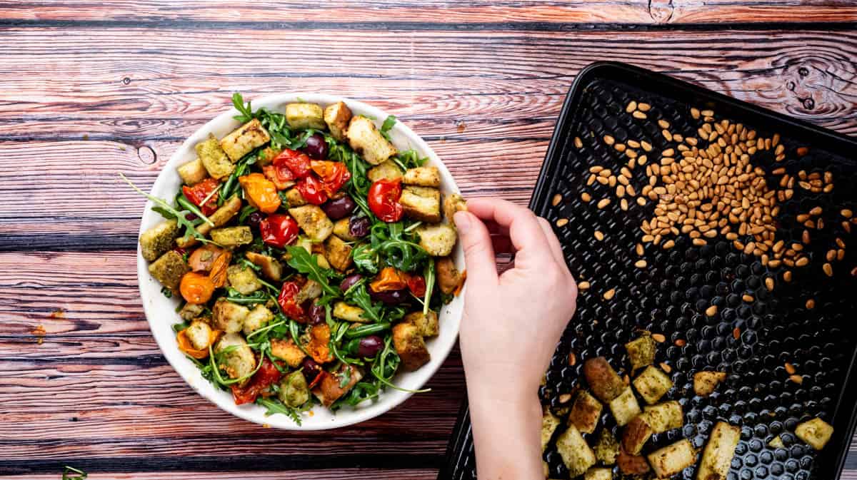 A hand is adding stale pesto bread cubes into the salad bowl.