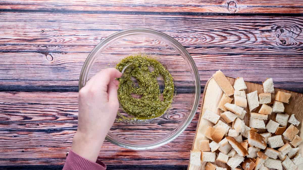 A hand is using a spoon to mix pesto and olive oil together in a glass bowl.