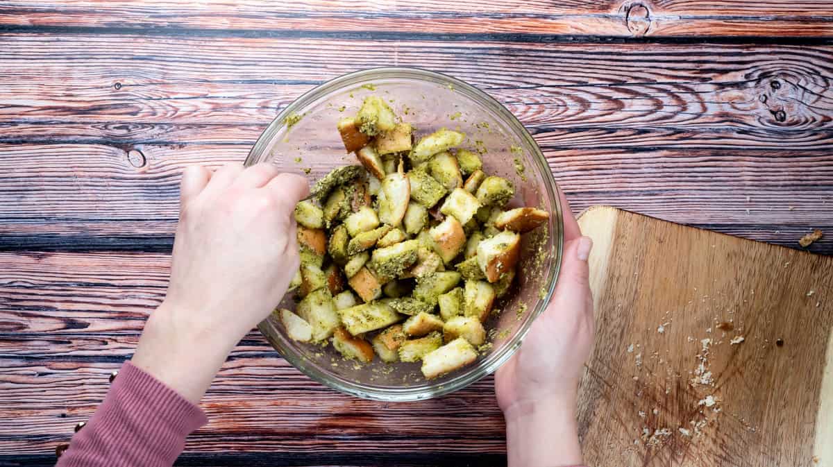 Two hands are stirring stale bread cubes into the pesto mixture to coat them.